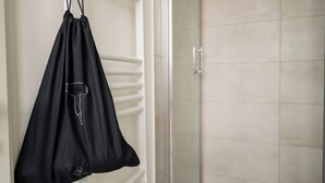 Un sac noir est suspendu à un mur blanc, et on aperçoit à l’arrière-plan une zone aménagée pour prendre une douche.