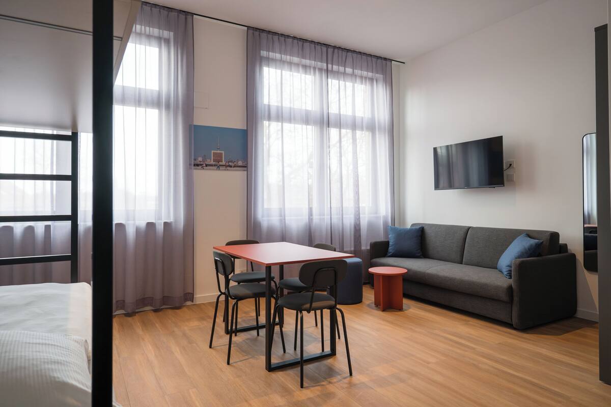 A modern living room with a sofa, a TV, a dining table with chairs, and a window with curtains.