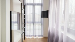 Une chambre avec un lit, un téléviseur fixé au mur et une fenêtre ornée de rideaux légers.