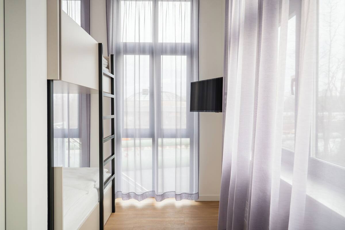 A room with a bed, a TV mounted on the wall, and a window with sheer curtains.