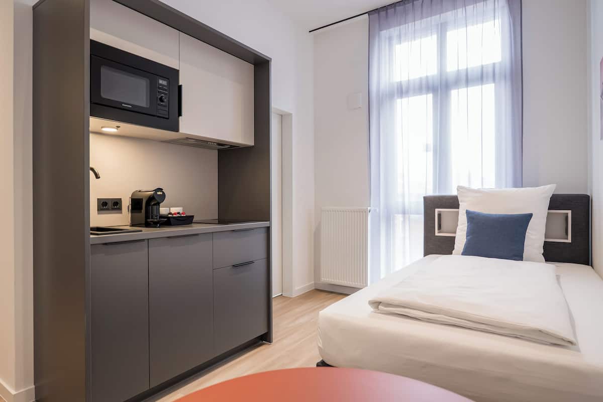 A compact hotel room with a kitchenette, a bed with a headboard, and a window with curtains.