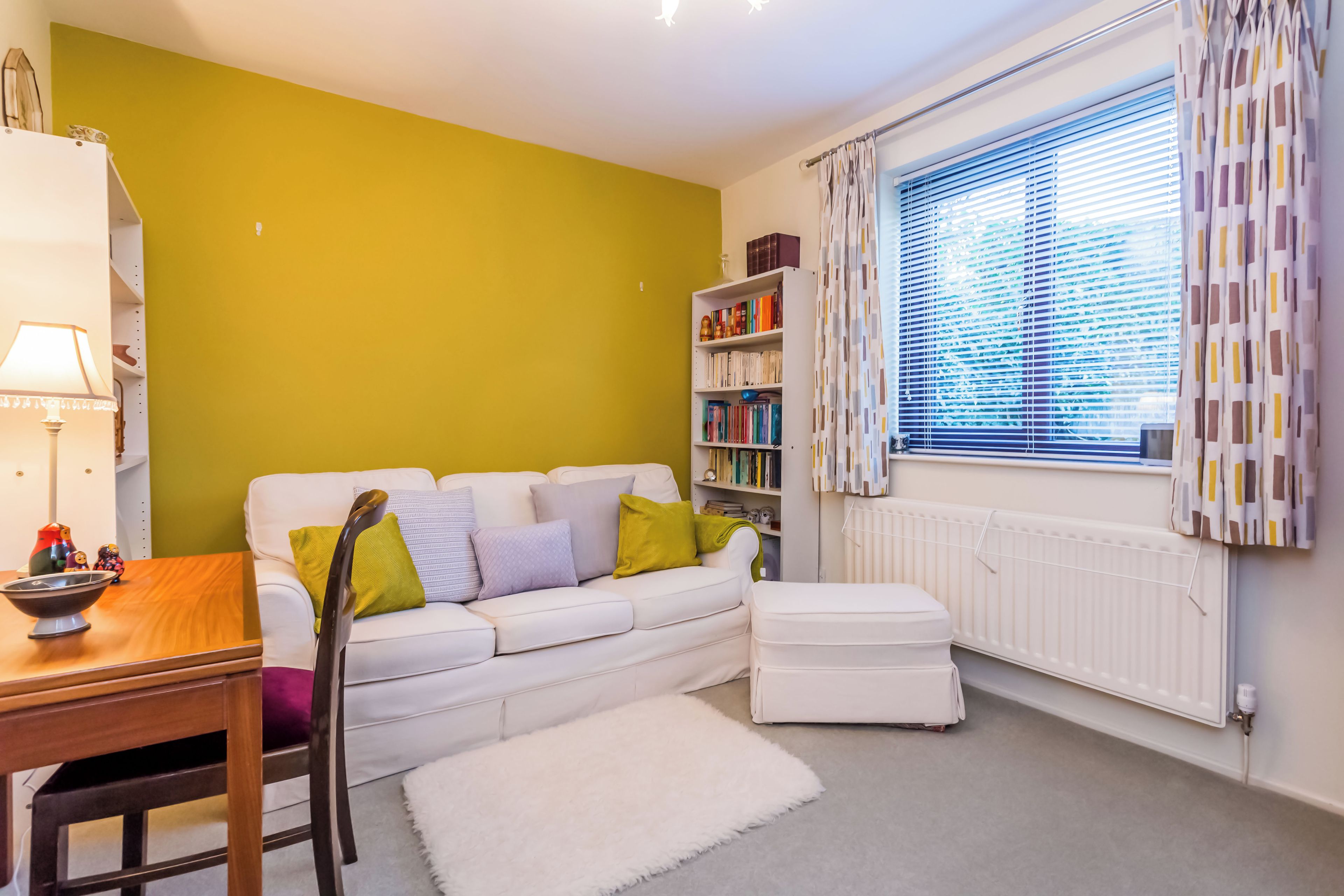 A cozy living room with a white sofa, a wooden table, a bookshelf, and a window with curtains.