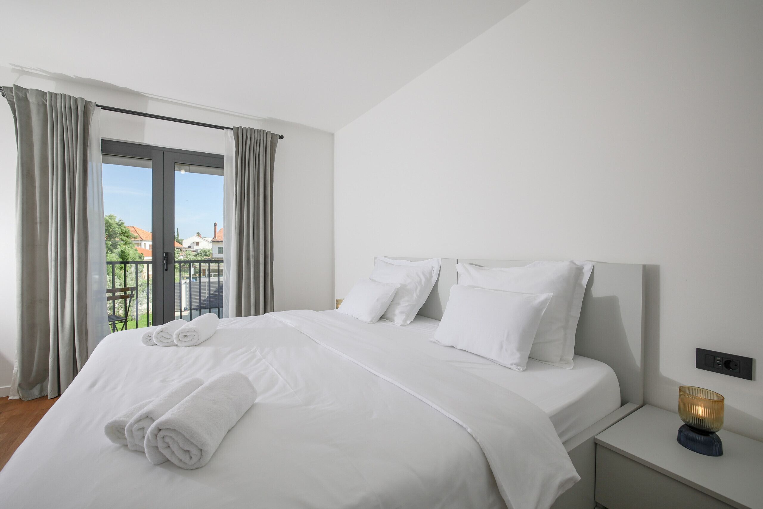 Un lit bien fait, avec du linge de lit blanc et des oreillers, une table de chevet avec une lampe, et une vue sur un balcon avec des rideaux.
