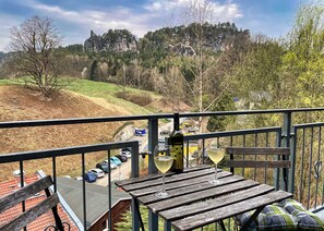 A wooden deck with a table and chairs, overlooking a parking area and a hilly landscape with trees and a rocky outcrop.