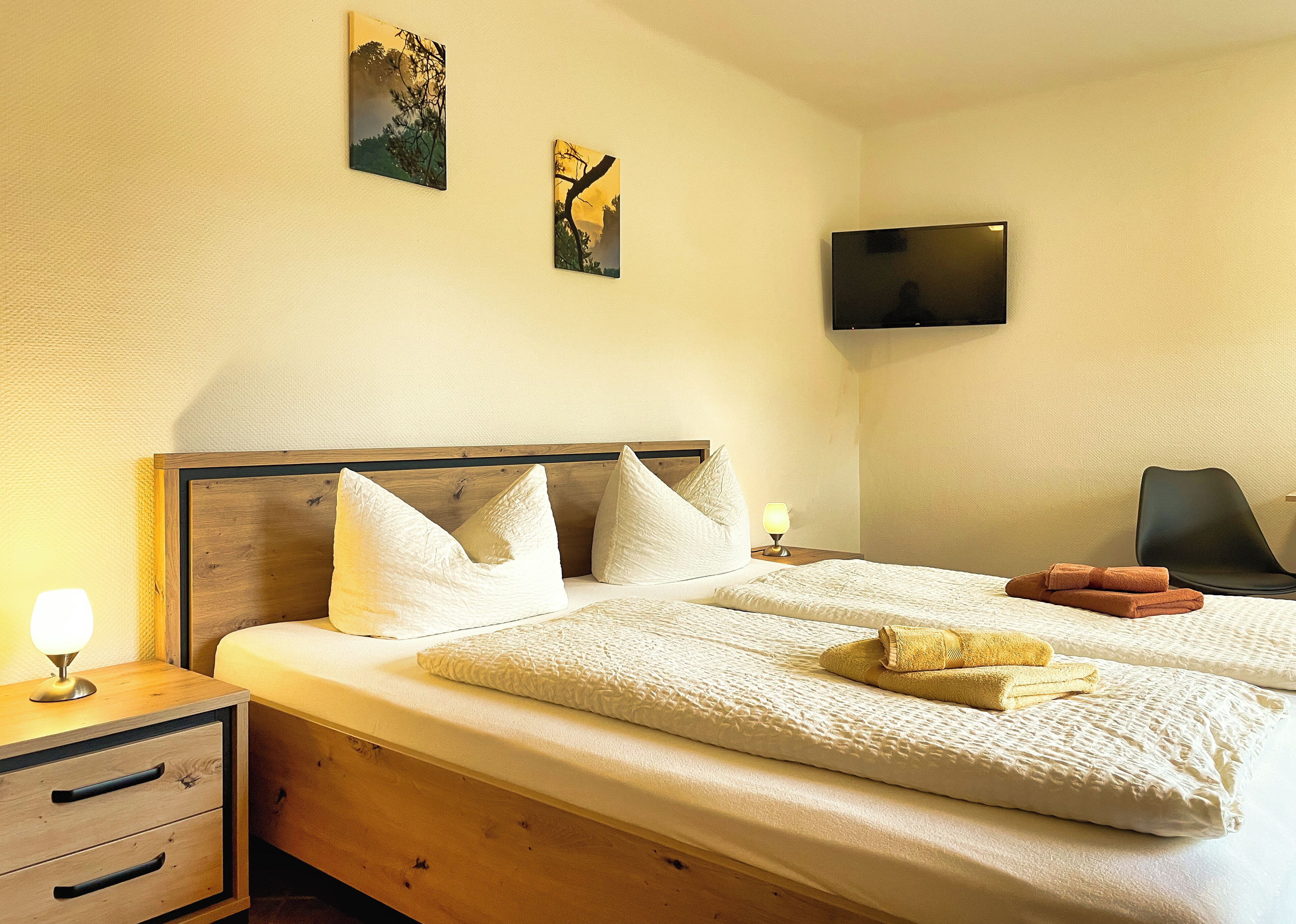 Ein Hotelzimmer mit Holz-Kopfteil, zwei Betten mit weißer Bettwäsche, einem Nachttisch mit Lampe, einem an der Wand montierten Fernseher und einem Sessel.
