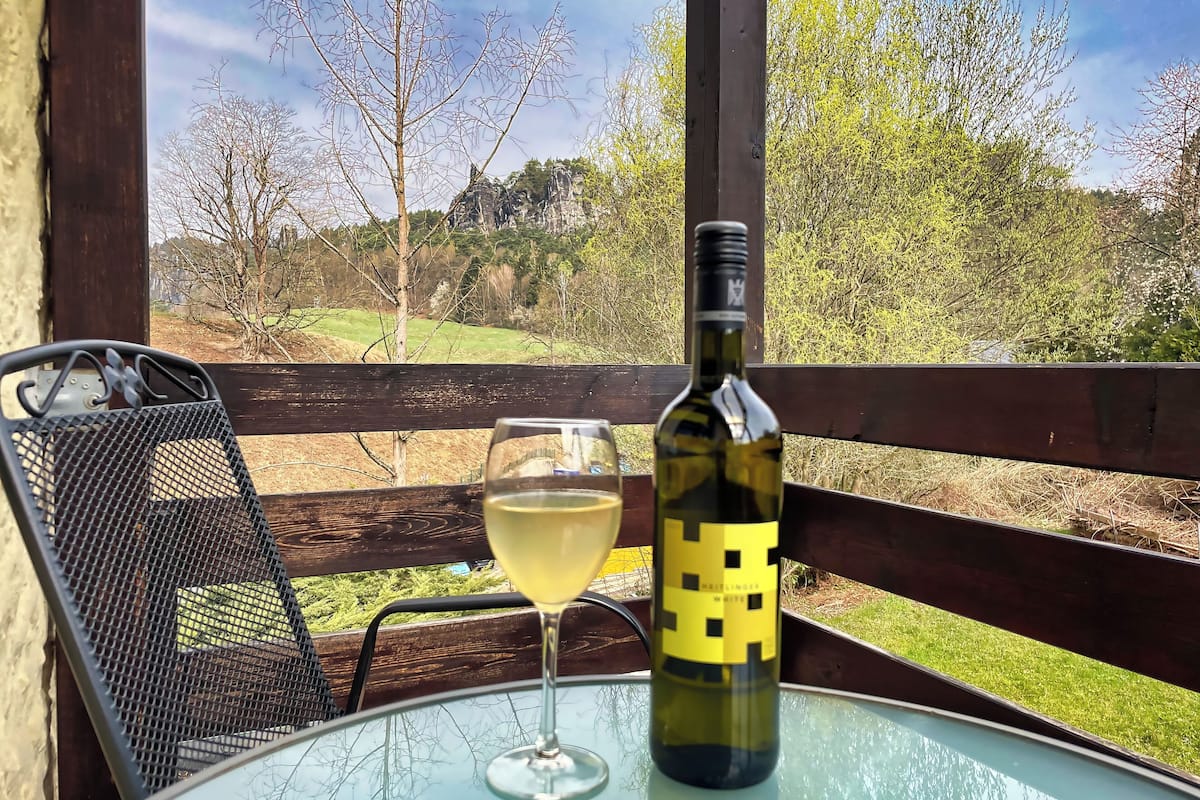 A bottle of wine and a glass on a table with a view of a grassy landscape and trees.