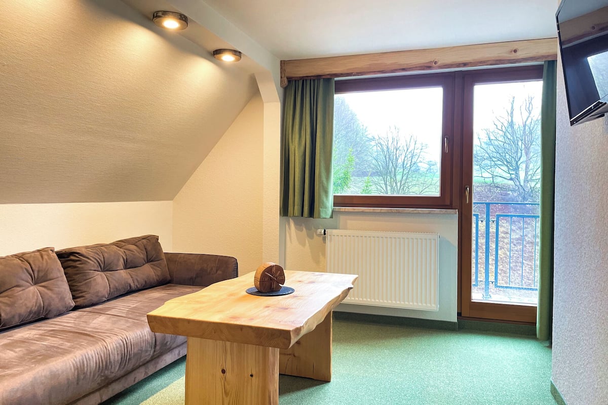 A room with a brown sofa, a wooden table with a clock, a window with curtains, and a radiator.
