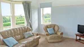 Una sala de estar con un sofá y un sillón de cuero color beige, un televisor sobre un mueble de madera y ventanas grandes con cortinas.