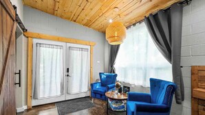 Una habitació amb sostre de fusta, dues grans finestres amb cortines, un sofà blau, una taula rodona petita i una làmpada penjant.