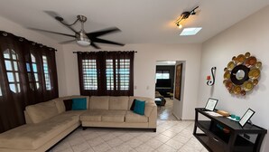 A living room with a beige sectional sofa, a ceiling fan, a flat-screen TV, and a wall-mounted decorative plate. - LaPlaya House 5 minutes from the best beaches! Ideal house for families (Manati)