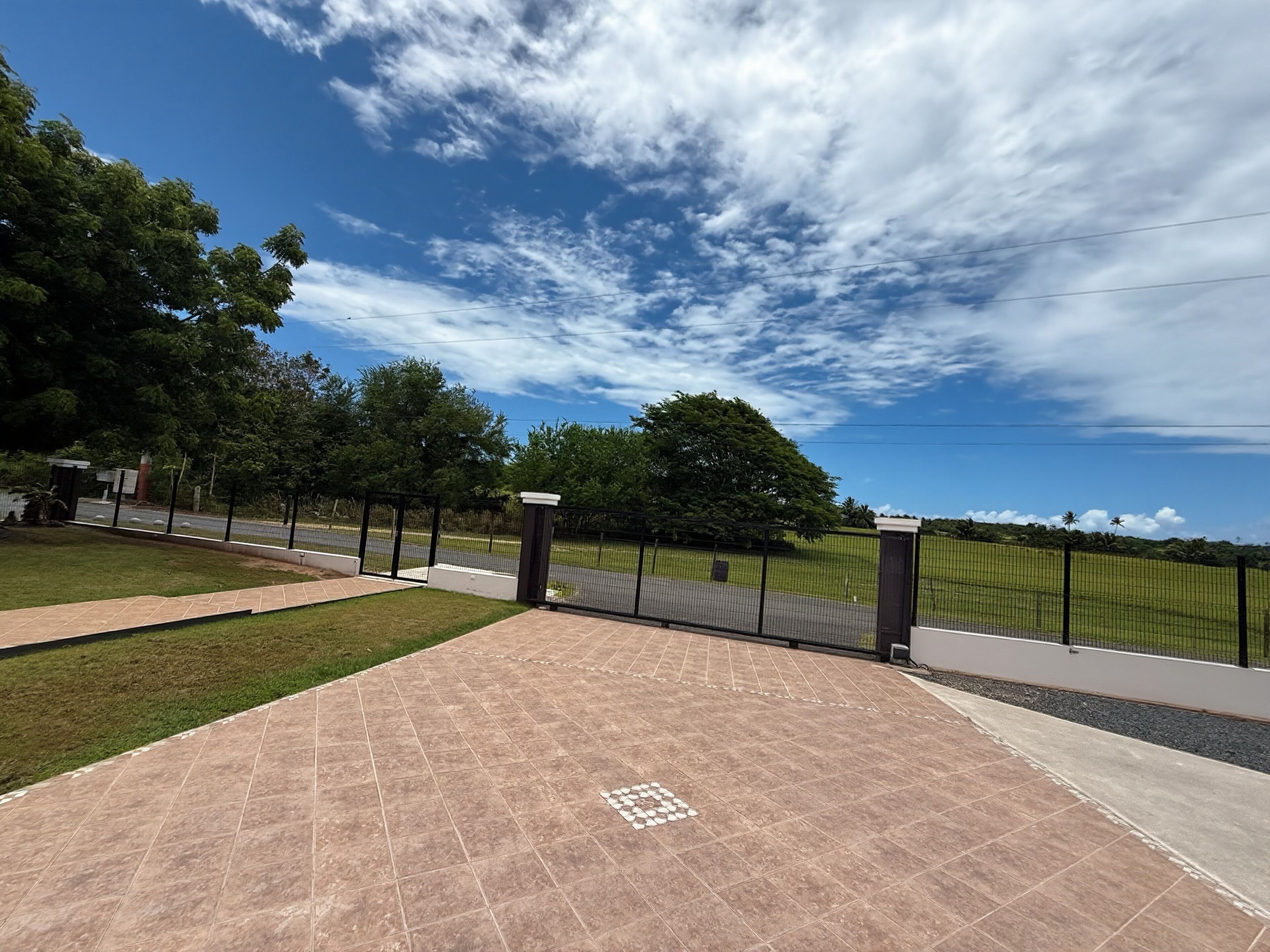 A paved outdoor area with a tiled floor, a black metal fence, and a grassy field with trees in the background under a partly cloudy sky.