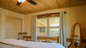 Een slaapkamer met een bed, houten plafond, raam met gordijnen, houten stoel en een spiegel.