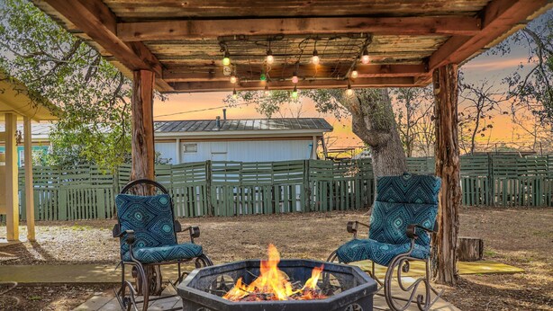 Een overdekt terras met een vuurkorf, twee gestreept stoelen en een houten pergola met lichtslingers.
