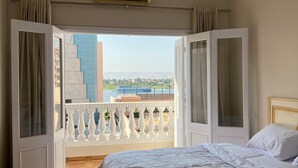Un dormitorio con una cama grande, una puerta abierta que da a un balcón con vistas a un río y edificios.