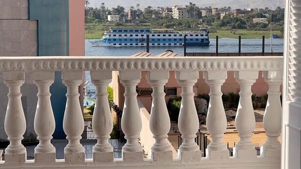 Un balcón con vista a un río, una embarcación y un paisaje urbano distante.