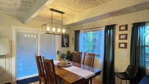 A dining area with a wooden table, chairs, and a chandelier. There is a window with curtains and a view of greenery outside.