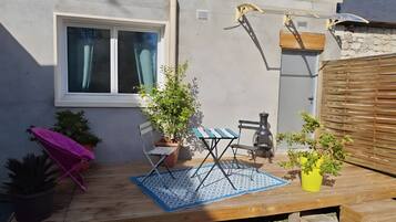 Eine Holzterrasse mit Tisch, Stühlen, einer Zimmerpflanze, einem Fenster und einer Wandleuchte.