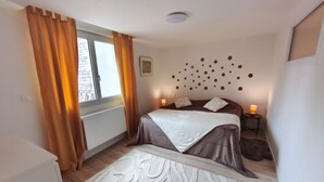 Un dormitorio con cama, ventana con cortinas, radiador, pared decorada con motivos circulares y mesita de noche con lámpara.
