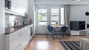 Une cuisine moderne avec des armoires blanches, un four encastré et un four à micro-ondes. On y trouve un coin repas avec une table et des chaises, ainsi qu’un téléviseur posé sur un meuble.
