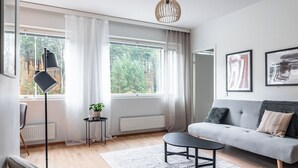 Un salon moderne avec un canapé gris, un lampadaire noir et une table basse ronde. Il y a deux fenêtres avec des rideaux légers, un radiateur et une plante en pot sur une petite table.