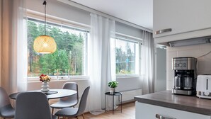 Une cuisine moderne équipée d’une machine à café, un coin repas avec une table et des chaises, et de grandes fenêtres offrant une vue sur les arbres.