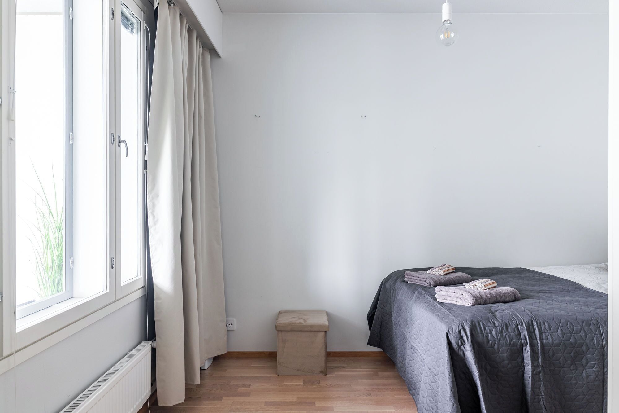 Une chambre à coucher avec un lit, un petit tabouret, un radiateur et une fenêtre avec des rideaux.