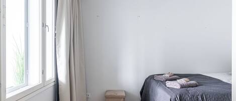 Un dormitorio con cama, un taburete pequeño, un radiador y una ventana con cortinas.
