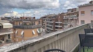 Een balkon met een betonnen balustrade, een zwarte metalen stoel en uitzicht op meerlaagse gebouwen met balkons en ramen.