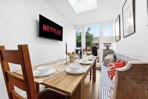 Ein Essbereich mit einem Holztisch, gedeckt für vier Personen, einem an der Wand befestigten Fernseher, der 'NETFLIX' zeigt, und einer Aussicht ins Freie durch die Fenster.