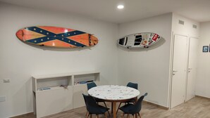 A room with a round table and four chairs, a white cabinet with shelves, and two surfboards on the wall. - Gisella Surf House (Ladispoli)