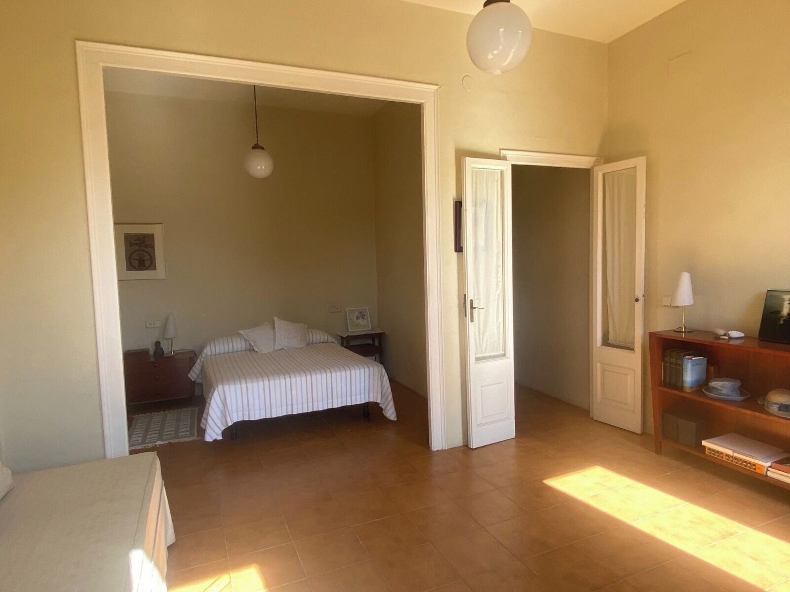 Quarto com cama, mesa de cabeceira, cômoda e porta que dá para outro ambiente.
