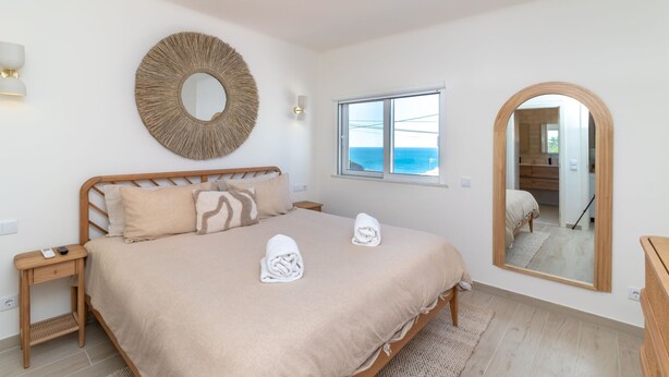 Ein Schlafzimmer mit einem Bett, einem runden Spiegel, einem Fenster mit Meerblick und einer Holzkommode.