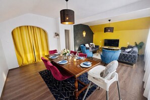 Un coin repas avec une table en bois dressée pour quatre personnes, des chaises rouges et blanches, un rideau jaune et une suspension noire.