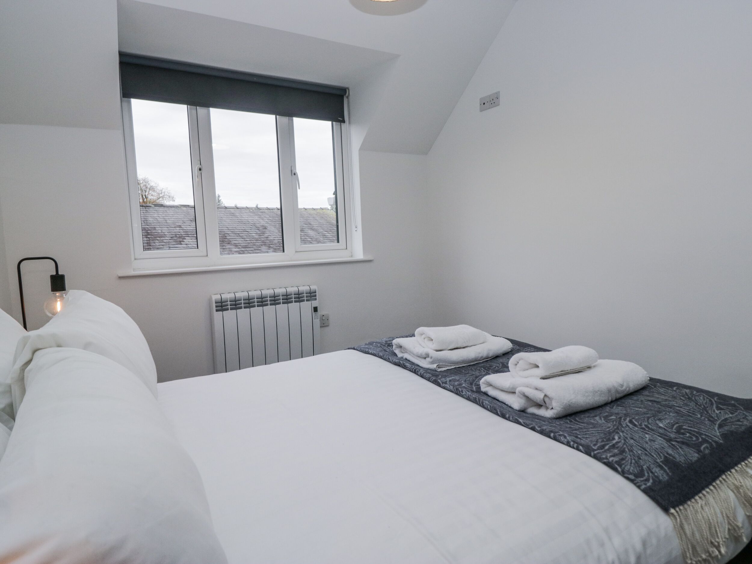 Un dormitorio con cama, radiador, ventana y lámpara de noche.