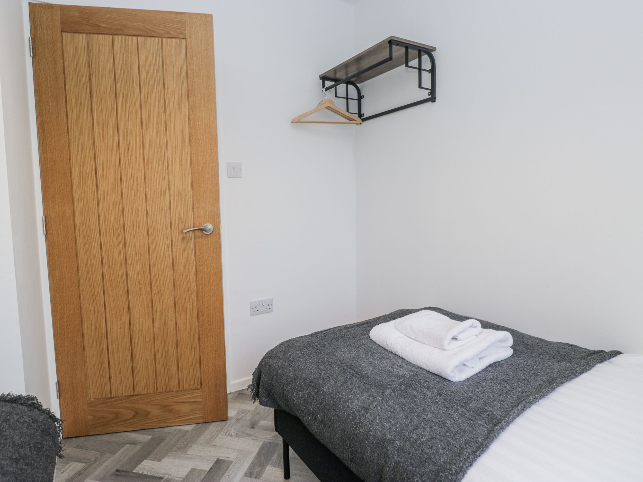 Ein Schlafzimmer mit Holztür, einem Bett mit grauer Decke und weißem Handtuch sowie einem an der Wand befestigten Regal mit Kleiderhaken.