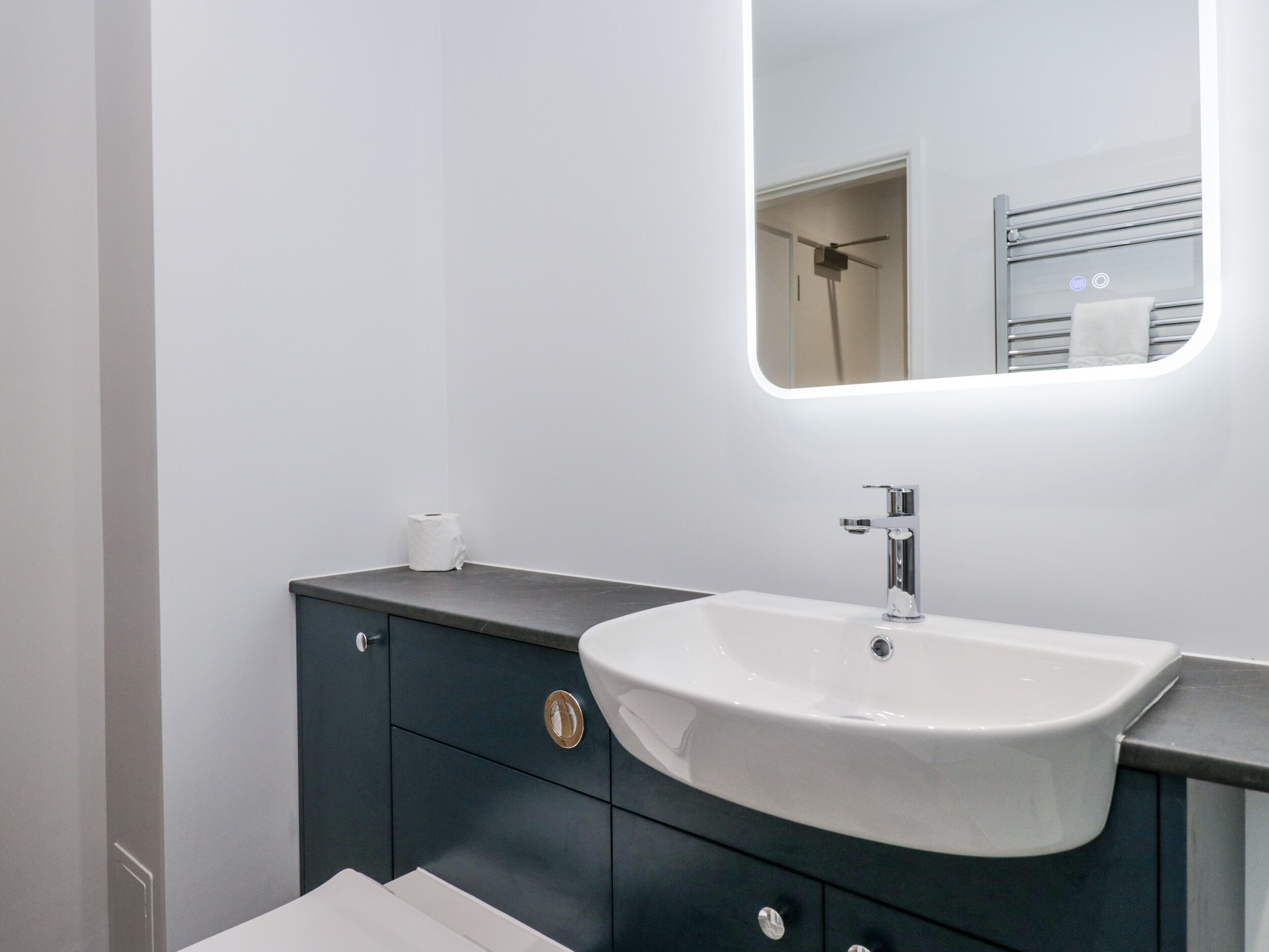 A modern bathroom with a white sink, dark cabinet, and a mirror with a lighted frame.