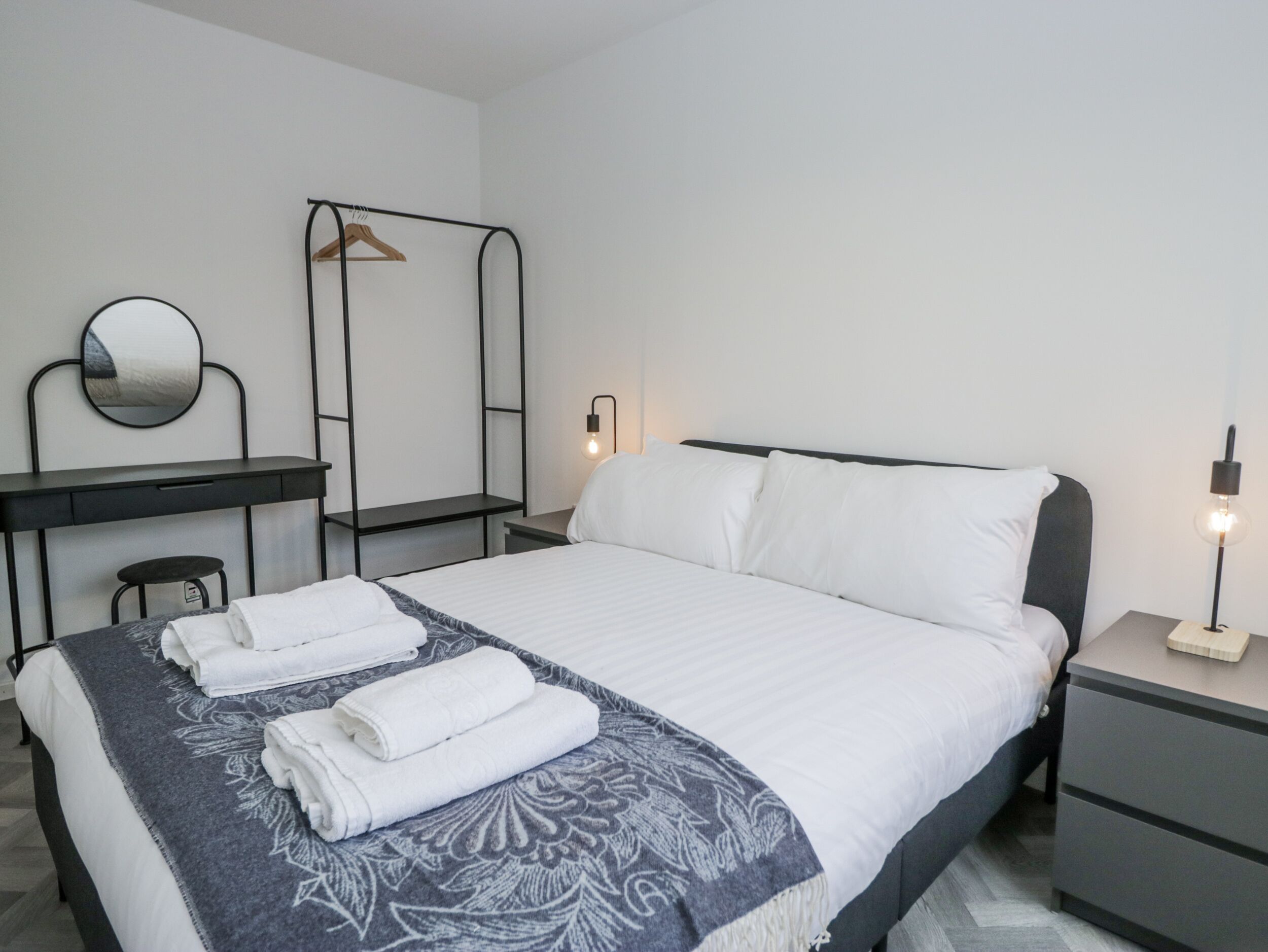 A neatly arranged bedroom with a bed, bedside tables, a desk, a mirror, and a hanging rack.