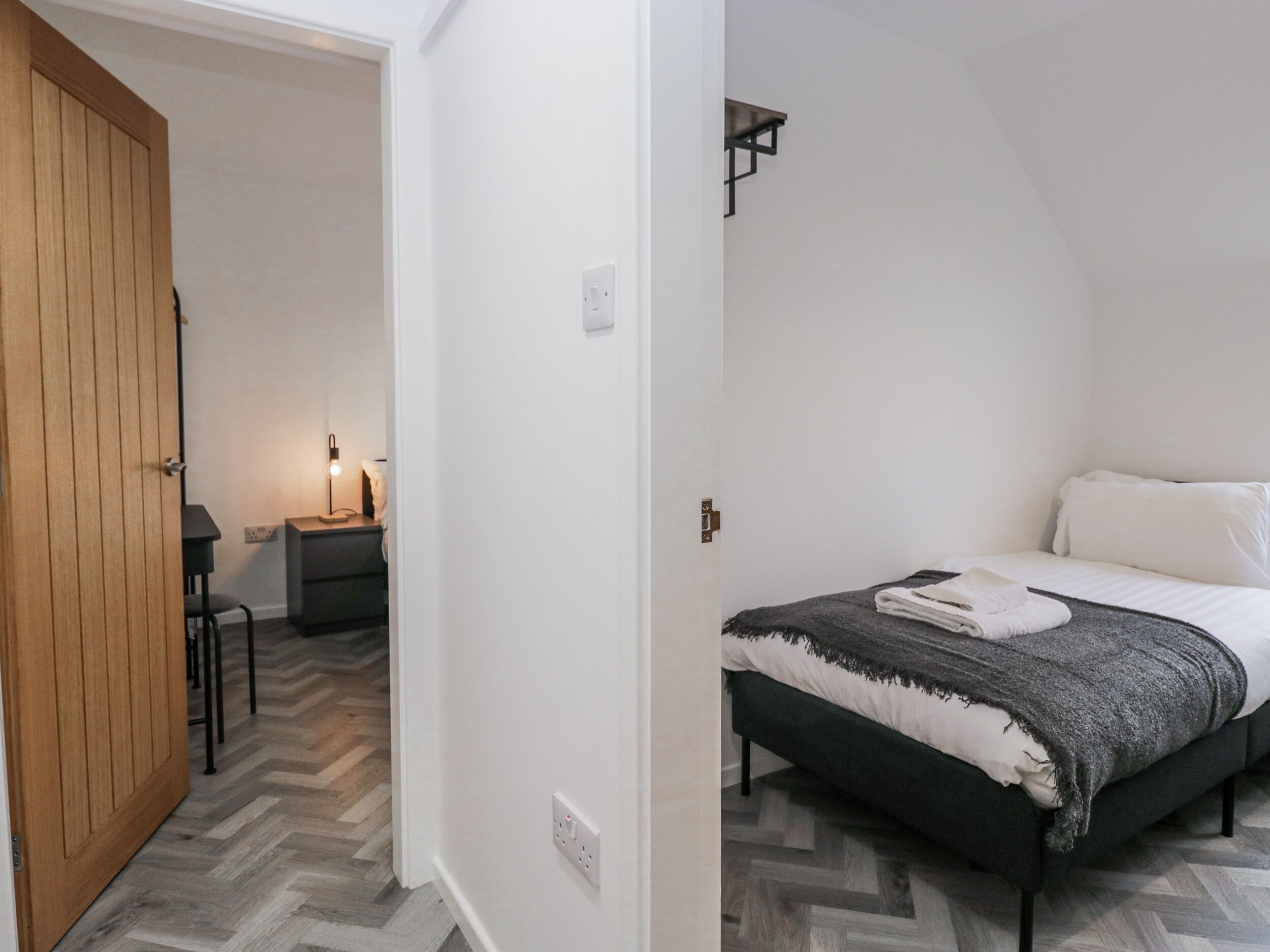 Quarto com cama, porta de madeira, mesa de cabeceira com abajur e piso com padrão herringbone.