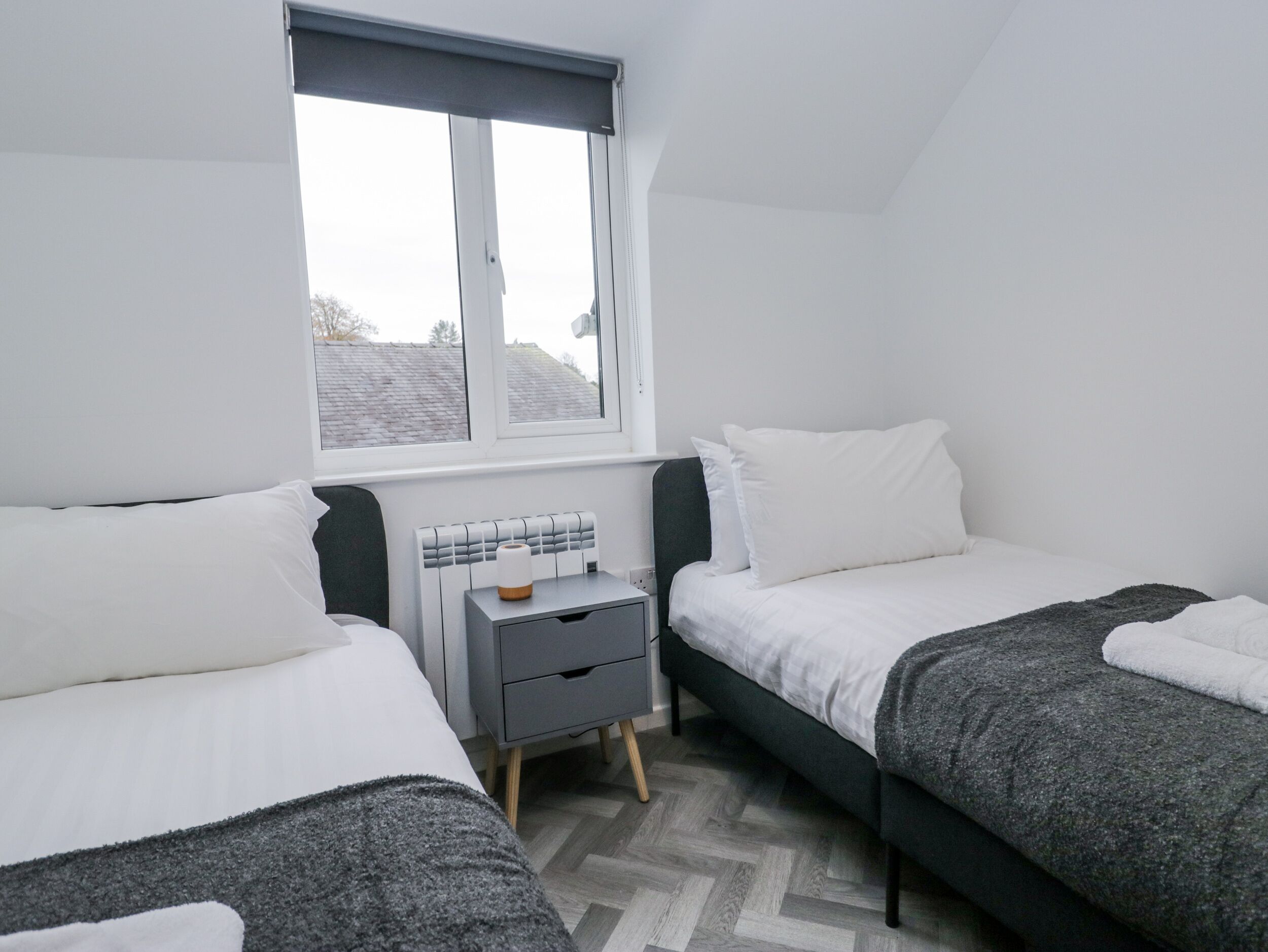 A bedroom with two beds, a window, a bedside table, and a radiator.
