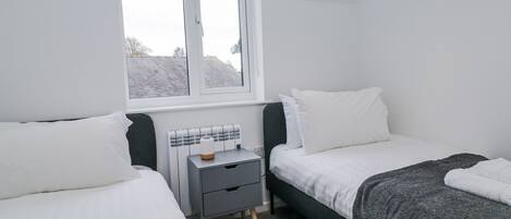 A bedroom with two beds, a window, a bedside table, and a radiator.
