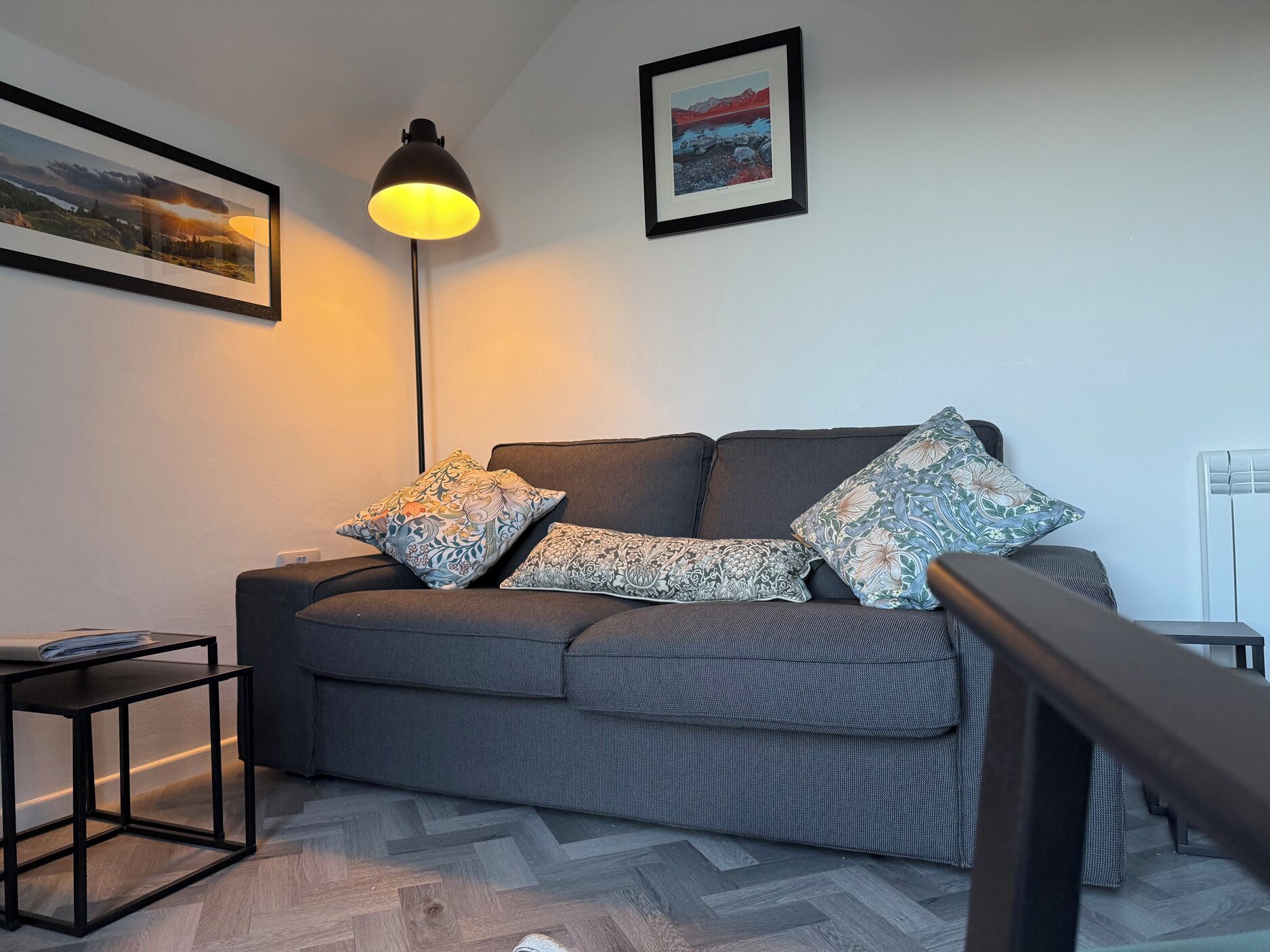 A modern living room with a grey sofa, patterned cushions, a black floor lamp, and a framed landscape painting on the wall.