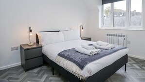 Ett modernt sovrum med en stor säng, två sängbord, ett element och ett fönster med utsikt över byggnader.