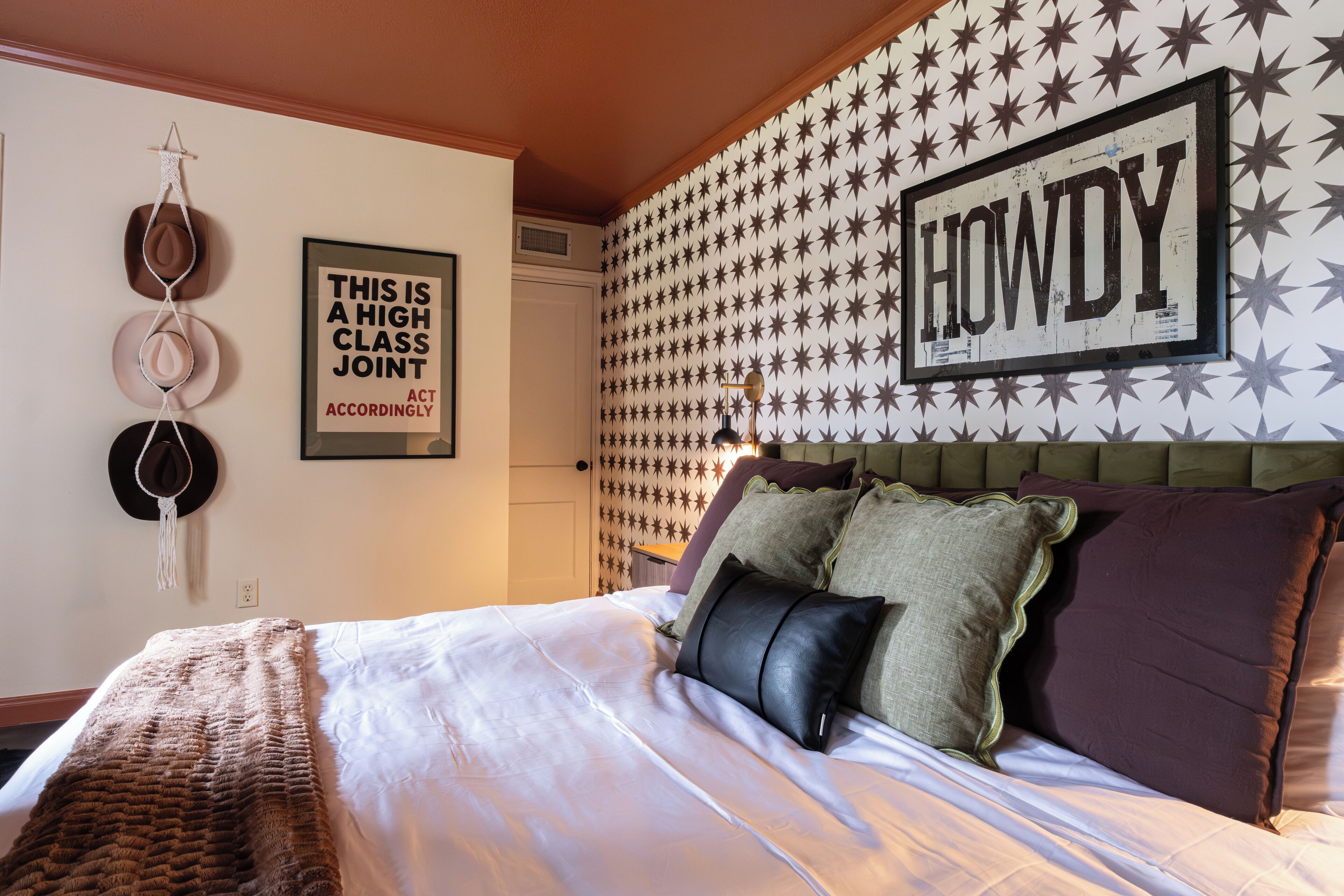 Una cama bien hecha con una colcha blanca y varias almohadas, una pared con un diseño de estrellas, y un letrero enmarcado que dice 'HOWDY'.