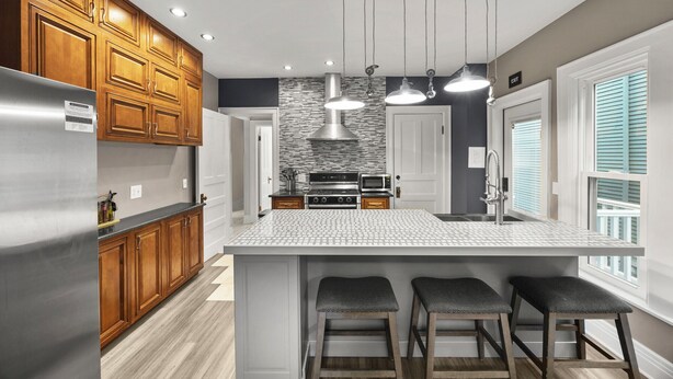 A modern kitchen with wooden cabinets, a stainless steel refrigerator, a central island with stools, and pendant lighting. - 3-bedroom apartment-Downtown, Modern, Central, Renovated & Cozy (Buffalo)