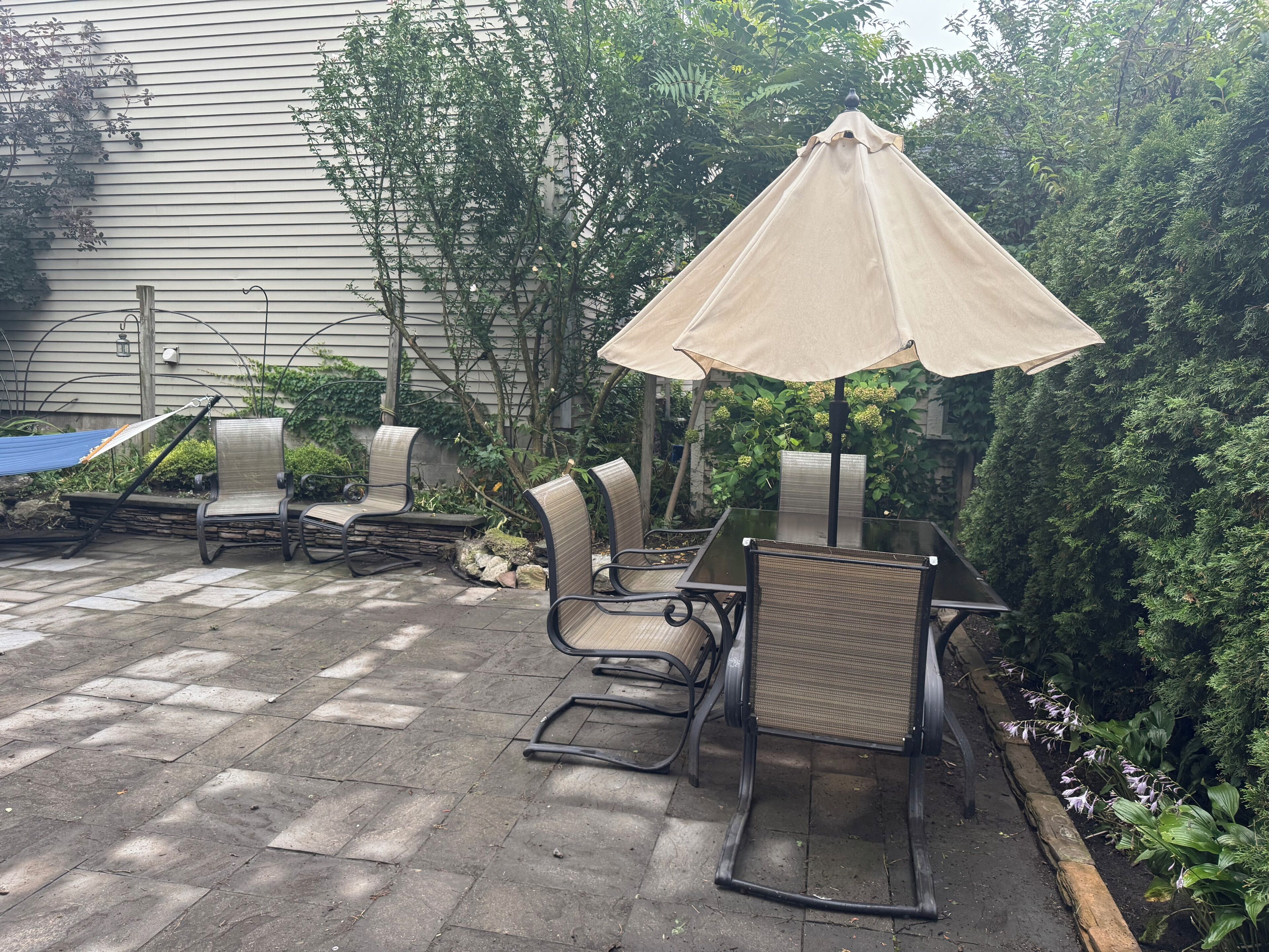 An outdoor patio with a table and chairs, surrounded by greenery and a house in the background.