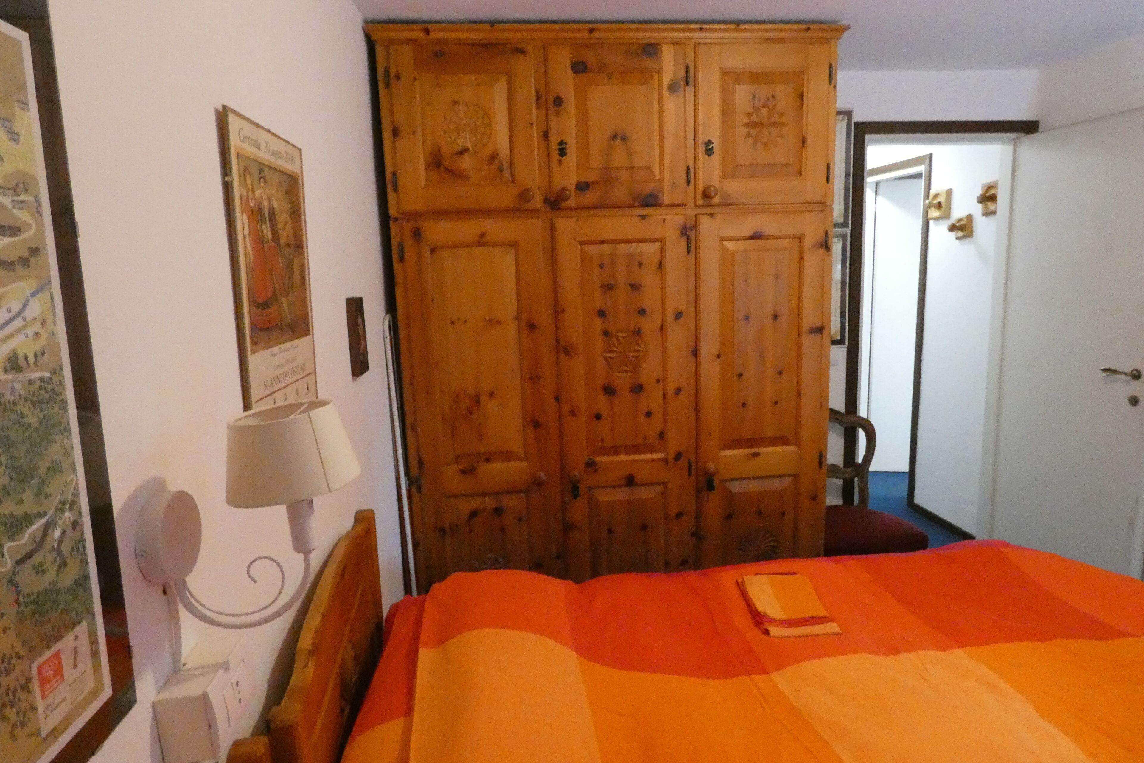 A bedroom with a large wooden wardrobe, a bed with an orange blanket, a wall-mounted lamp, and a framed picture.