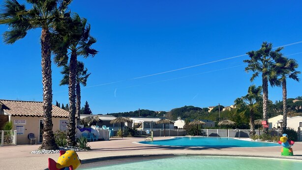 Zona de piscina con palmeras, cielo azul despejado y un cartel que indica un puesto de primeros auxilios.