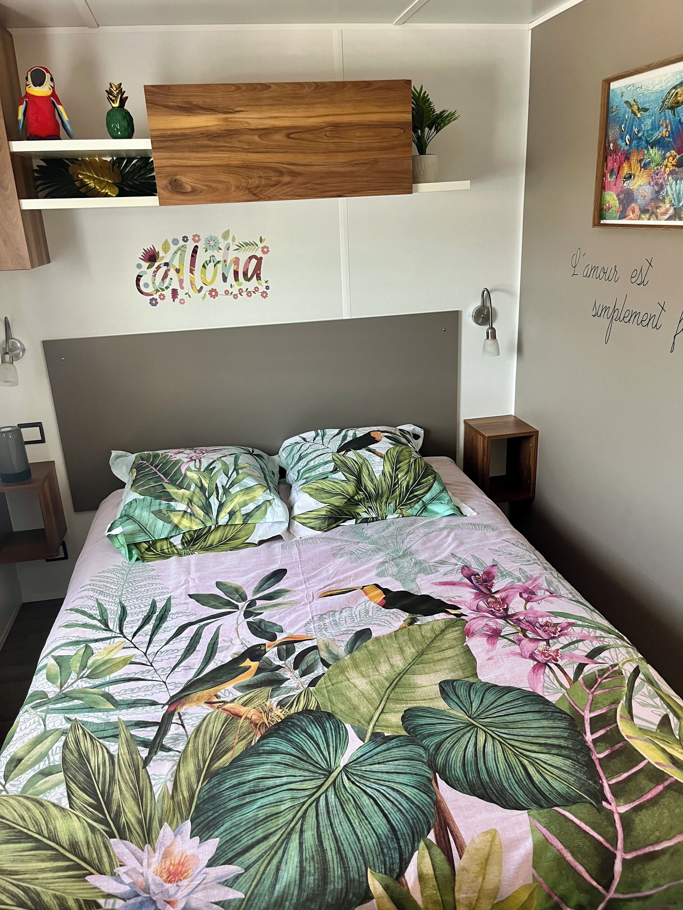 A bedroom with a bed featuring tropical leaf and bird prints, a wooden shelf with decorative items, and a wall with a floral decal and a French phrase.