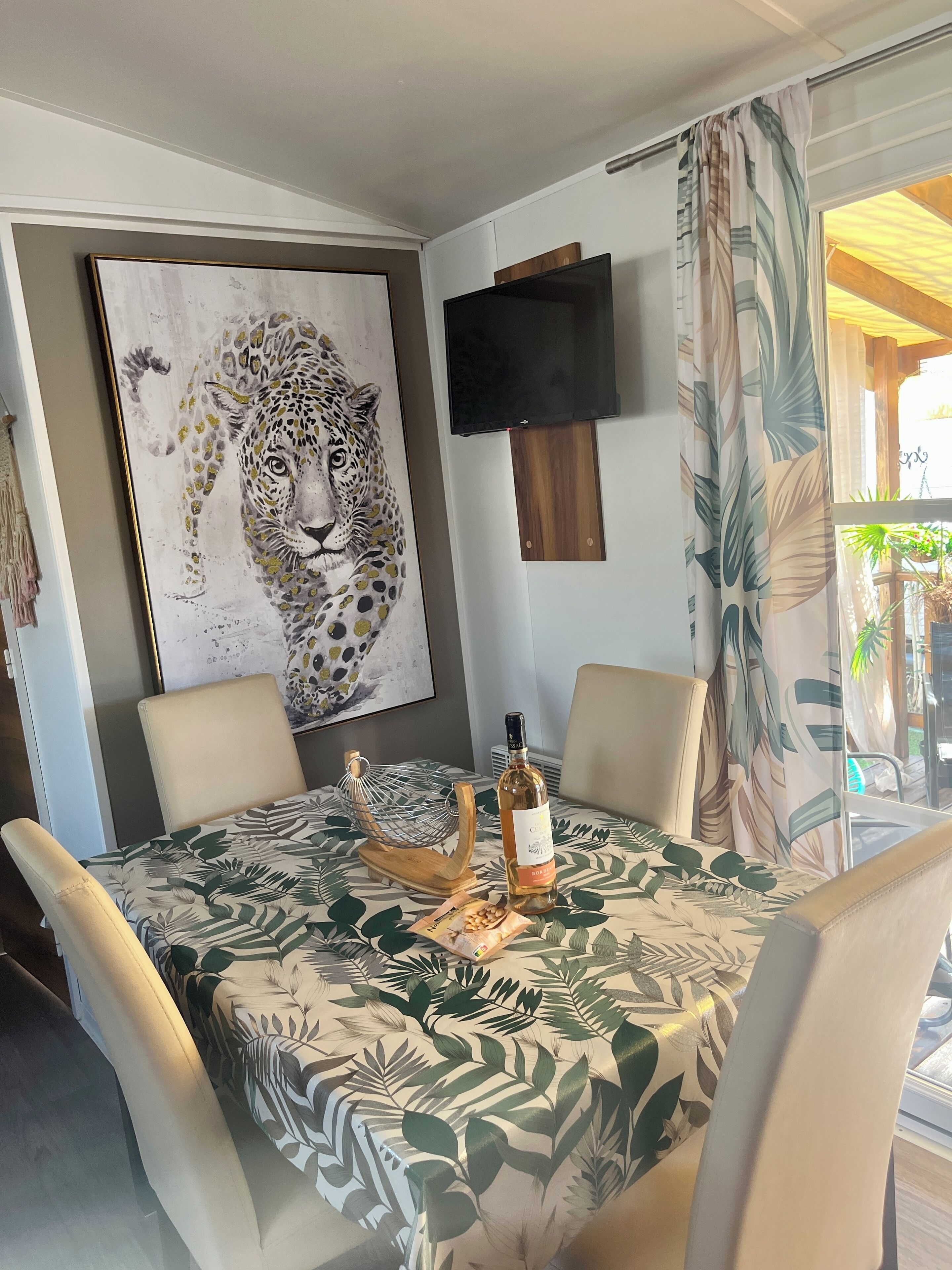 Un coin repas avec une table recouverte d’une nappe à motifs de feuilles, entourée de chaises. Une bouteille de vin et un élément décoratif ornent la table. Un grand tableau représentant un léopard est accroché au mur au-dessus de la table.