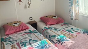 Un dormitorio con dos camas, cada una con una colcha estampada en rosa y blanco, un estante de madera arriba y una pared decorada con pegatinas de monos y pájaros.